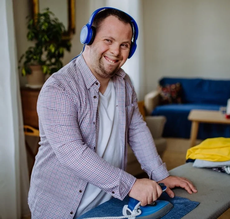 Man with down syndrome doing ironing in living room with headphones on and smiling at camera