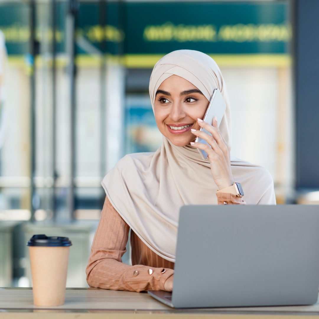 Smiling friendly cute millennial arab muslim woman in hijab speaks on phone and types on laptop in